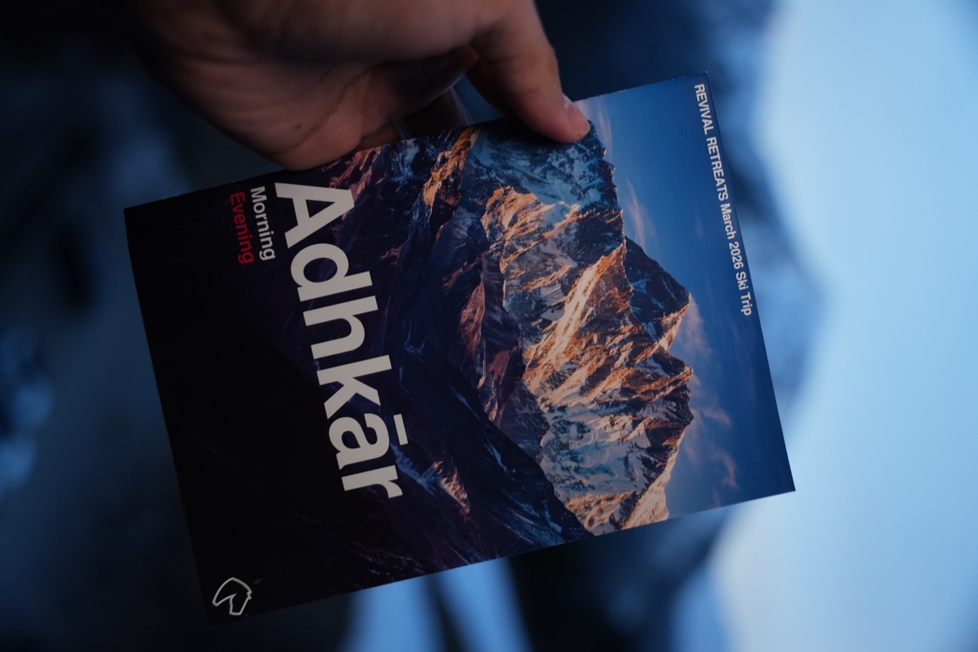 A hand holding the Revival Retreats Adhkār booklet against mountains at dusk